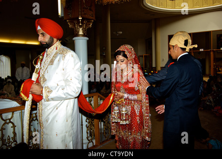 Traditional East Indian wedding groom wearing bright red turban leading bride around Sikh temple altar joined marriage scarf Stock Photo