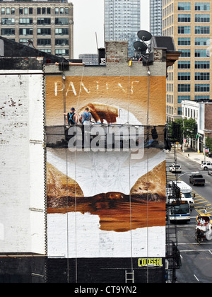 2 sign painters on scaffold work on hair products billboard painted on side of old brick tenement in Hells Kitchen New York City Stock Photo
