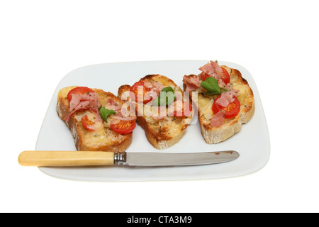 Toasted ciabata bread topped with cheese,tomato, ham and basil on a plate with a knife isolated against white Stock Photo