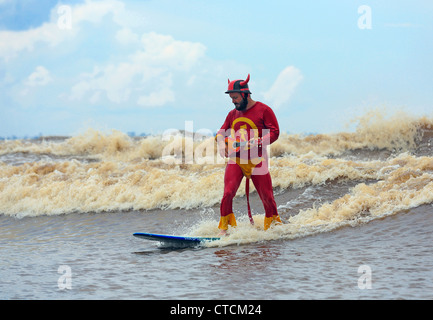 Strange man in red superhero costume river surfing a wave while playing a ukulele, on the wave called Bono or the 7 Ghosts. Stock Photo