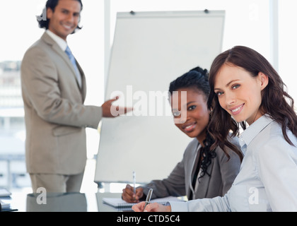 Two young smiling employees sitting at the desk with a colleague behind them Stock Photo
