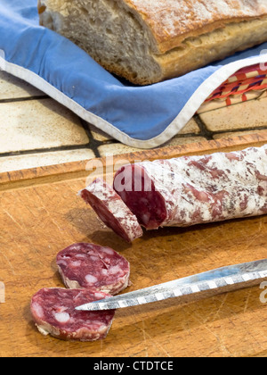 French dry sausage on a wooden cutting board with slices cut off and a knife. A bread is present in background. Stock Photo