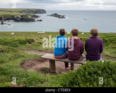 Three people sitting on a bench facing out to sea at the top of cliffs at Porthcothan Bay near Padstow Cornwall UK Stock Photo