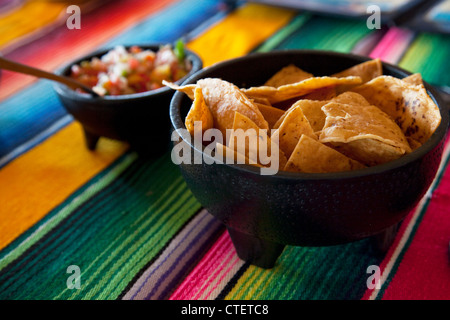 Colourful Mexican table cloth with a bowl of taco chips and fresh salsa, served in traditional black Mexican bowls Stock Photo