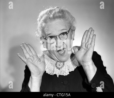 1960s ELDERLY WOMAN IN GRANNY GLASSES HOLDING HAND UP NEAR FACE WITH MOUTH OPEN LOOKING AT CAMERA Stock Photo
