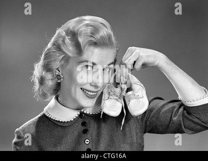 1950s 1960s SMILING BLOND WOMAN COMMUNICATING THAT SHE IS MOTHER TO BE BY HOLDING UP A PAIR OF BABY SHOES LOOKING AT CAMERA Stock Photo