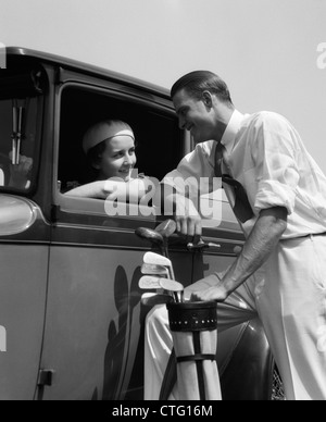 1930s WOMAN IN DRIVER'S SEAT OF CAR LEANING OUT OF WINDOW TALKING TO MAN STANDING HOLDING GOLF BAG Stock Photo