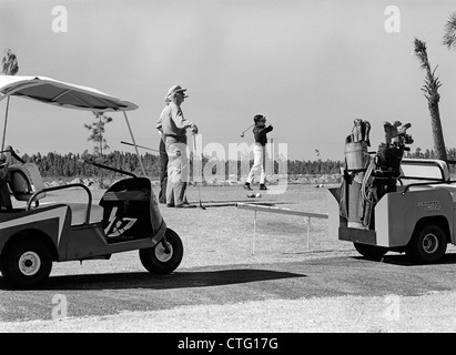 1960s THREE MEN PLAYING GOLF TEEING OFF FROM TEE WITH DRIVER NEAR GOLF CARTS OUTDOOR Stock Photo
