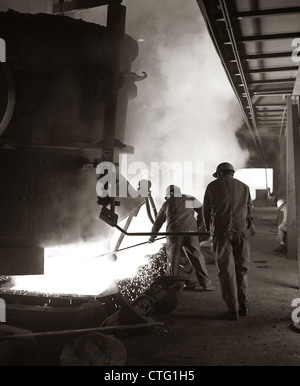 1960s PAIR OF STEEL WORKERS WEARING HARD HATS STIRRING & STOKING MOLTEN METAL POURING FROM CRUCIBLE Stock Photo