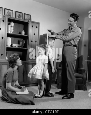 1960s FATHER MEASURING DAUGHTER & SON BACK-TO-BACK WITH MOTHER RECORDING INFORMATION Stock Photo