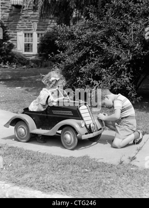 1940s 1930s BOY ON SIDEWALK FIXING HEADLIGHT OF TOY CAR DRIVEN BY LITTLE GIRL PLAYING OUTDOOR Stock Photo