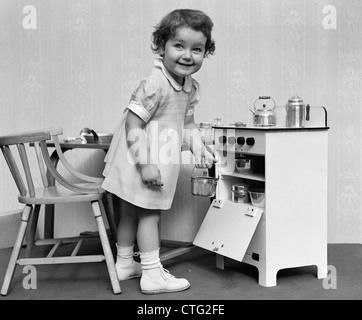 1940s 1950s SMILING LITTLE GIRL TAKING TOY POT OUT OF OVEN IN MINIATURE TOY COOKING STOVE Stock Photo