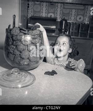 1950s WIDE-EYED GIRL WITH MOUTH AGAPE REACHING UP INTO COOKIE JAR TAKING ANOTHER CHOCOLATE CHIP COOKIE Stock Photo