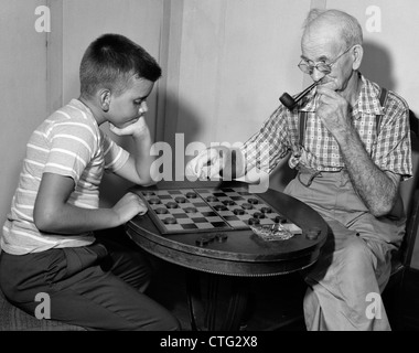 1950s BOY PLAYING CHECKERS WITH GRANDFATHER SMOKING PIPE & PICKING UP PIECE TO JUMP HIS CHECKER Stock Photo