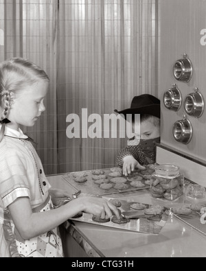 1960s BLOND GIRL TAKING COOKIES OFF TRAY AT KITCHEN COUNTER YOUNGER BOY WITH COWBOY HAT BANDANA MASK REACHES TO STEAL THEM Stock Photo