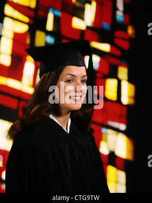 1970s SMILING BRUNETTE GRADUATE GIRL WOMAN WEARING ROBE MORTARBOARD STAINED GLASS BACKGROUND Stock Photo