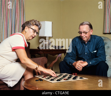 1970s SENIOR COUPLE MAN AND WOMAN SITTING IN LIVING ROOM PLAYING CHECKERS Stock Photo