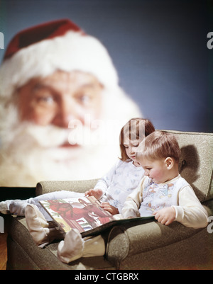 1970s CHILDREN READING CHRISTMAS STORY BOOK SANTA CLAUS IN BACKGROUND Stock Photo