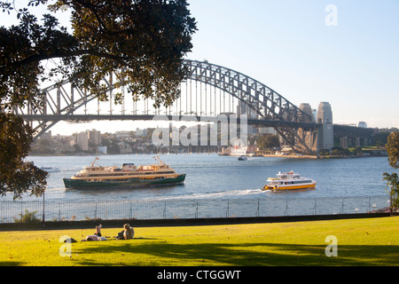 Two women lying in park at sunset with Manly Ferry and boat passing and Sydney Harbour Bridge in background Sydney NSW Australia Stock Photo