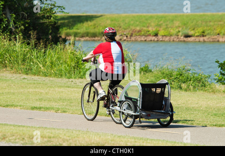 An obese woman , pulling a kiddy cart, rides her bicycle on trails by a lake in Oklahoma, USA.
