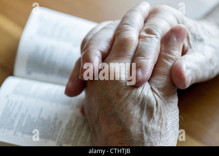 weathered old man's hands clasped in prayer over open Bible Stock Photo