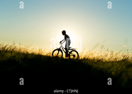 Boy riding a bicycle through grass at sunset. Silhouette. UK Stock Photo