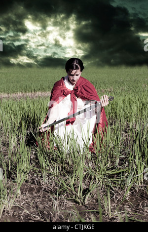 a woman in a red cloak sitting on a field with a sword Stock Photo