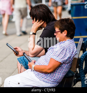 a man woman couple reading e-books on their Amazon Kindle readers, sitting outside on a warm day, UK Stock Photo