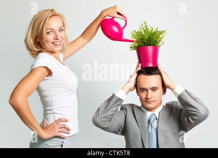 Serious businessman holding a plant on his head, cheerful young woman watering it Stock Photo