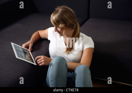 Young woman is using an IPad tablet computer, at home, on a sofa, couch. Surfing the Internet, using a wireless connection. Stock Photo