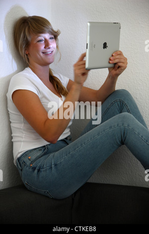 Young woman is using an IPad tablet computer, at home, on a sofa, couch. Surfing the Internet, using a wireless connection. Stock Photo
