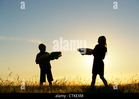 Boy and girl shooting each other with water guns at sunset. Silhouette. UK Stock Photo