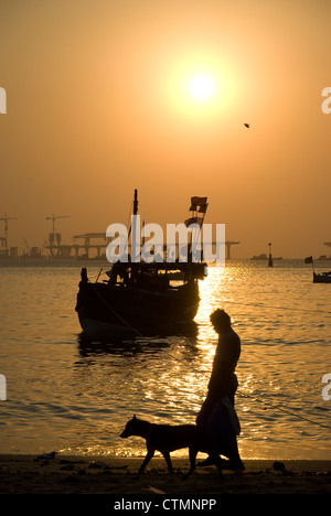 Man and dog walking on beach with a fishing boat and sealink in the background - Mumbai Stock Photo