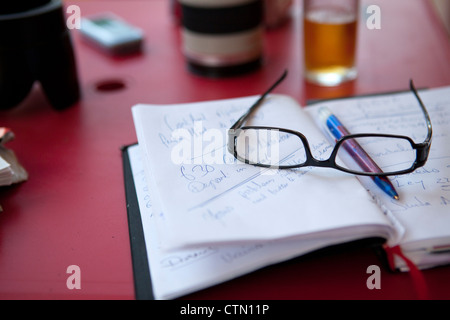 Red table top with open notebook  with pen and glasses on top, half a glass of beer and camera lens behind out of focus Stock Photo