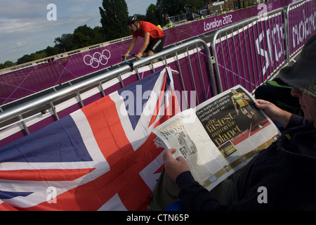 Spectator reads a tablid story written by Kamacain sprinter Eusain Bolt as a cyclist passes by before racers arrive on the first day of competition of the London 2012 Olympic 250km mens' road race. Starting from central London and passing the capital's famous landmarks before heading out into rural England to the gruelling Box Hill in the county of Surrey. Local southwest Londoners lined the route hoping for British favourite Mark Cavendish to win Team GB first medal but were eventually disappointed when Kazakhstan's Alexandre Vinokourov eventually won gold. Stock Photo