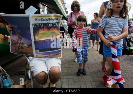 Local children walk past as a reader of the Daily Telegraph newspaper reads about the previous night's Olympic opening ceremony, on the first day of competition of the London 2012 Olympic 250km mens' road race. Starting from central London and passing the capital's famous landmarks before heading out into rural England to the gruelling Box Hill in the county of Surrey. Local southwest Londoners lined the route hoping for British favourite Mark Cavendish to win Team GB first medal but were eventually disappointed when Kazakhstan's Alexandre Vinokourov eventually won gold. Stock Photo