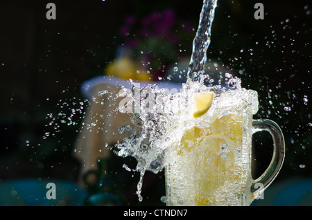 Water pouring and splahing into glass jug filled with lemons on sunny day. Stock Photo