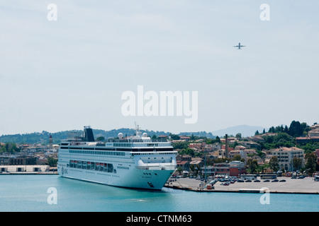 An airliner taking off from Corfu Airport passes over the cruise liner MSC Armonia, moored in Kerkyra port, Corfu Stock Photo