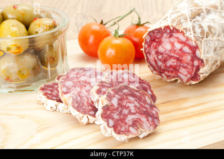 A dry cured salami sausage on a wooden cutting board with vine tomato's and olives in the background, shallow depth of field - s Stock Photo