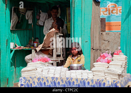 Hairdresser cutting hair in barbershop and child behind display selling goods in Govardhan, Uttar Pradesh, India Stock Photo