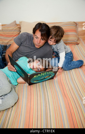 A father, daughter and son are lying on the bed reading a book together. Stock Photo