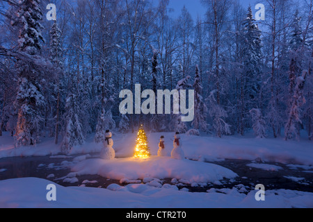 Snowman family  standing next Christmas tree on snow covered island in middle small stream in hoarfrost covered forest twilight Stock Photo