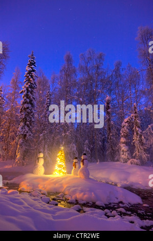 Snowman family  standing next Christmas tree on snow covered island in middle small stream in hoarfrost covered forest twilight Stock Photo