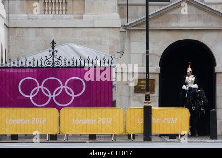 Olympic rings on a banner outside Horse Guards where a soldier from the Household Cavalry sits motionless on his horse during the London 2012 Olympics. Wrought iron railings are seen behind the banner at the sports venue hosting the volleyball in the centre of Westminster where governmental offices are located. The British Household Cavalry is classed as a corps in its own right, and consists of two regiments: Life Guards (British Army) and the Blues and Royals (Royal Horse Guards and 1st Dragoons). They are the senior regular regiments in the British Army, with traditions dating from 1660. Stock Photo