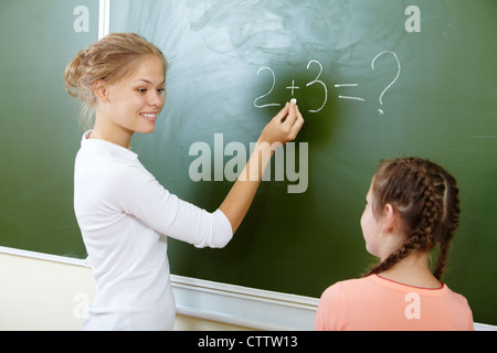 Photo of teacher writing sum on blackboard and looking at pretty pupil near by Stock Photo