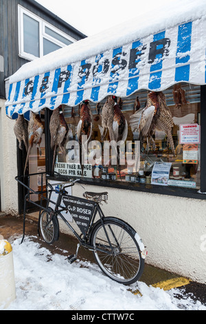 Village butcher's sop window and display in snow with pheasant hanging outside and delivery bike. Stock Photo