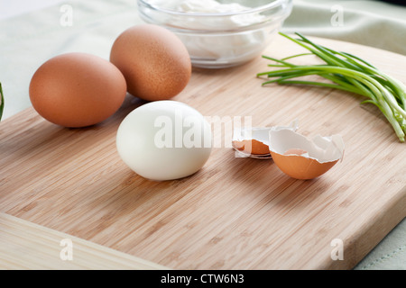 Peeled hard boiled egg with shells, two whole eggs and chives on cutting board. Stock Photo