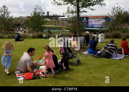 Families and spectators watch a televised rowing race on the grass in the Olympic Park during the London 2012 Olympics. The planting of 4,000 trees, 300,000 wetland plants and more than 150,000 perennial plants plus nectar-rich wildflower make for a colourful setting for the Games. This land was transformed to become a 2.5 Sq Km sporting complex, once industrial businesses and now the venue of eight venues including the main arena, Aquatics Centre and Velodrome plus the athletes' Olympic Village. After the Olympics, the park is to be known as Queen Elizabeth Olympic Park. Stock Photo