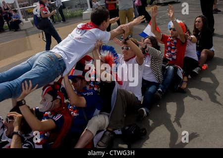 French spectators enjoy the spirit of the Olympics and international competition in the Olympic Park during the London 2012 Olympics. The sports fans support the weight of their friend and pass him overhead. This land was transformed to become a 2.5 Sq Km sporting complex, once industrial businesses and now the venue of eight venues including the main arena, Aquatics Centre and Velodrome plus the athletes' Olympic Village. After the Olympics, the park is to be known as Queen Elizabeth Olympic Park. Stock Photo