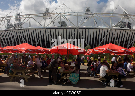 Spectators eat beneath Coca-Cola branded sponsor brolleys in near the main stadium in the Olympic Park during the London 2012 Olympics. Coca-Cola Company has supported the Olympic Games began in 1928, now a 92 years association without interruption. This land was transformed to become a 2.5 Sq Km sporting complex, once industrial businesses and now the venue of eight venues including the main arena, Aquatics Centre and Velodrome plus the athletes' Olympic Village. Stock Photo